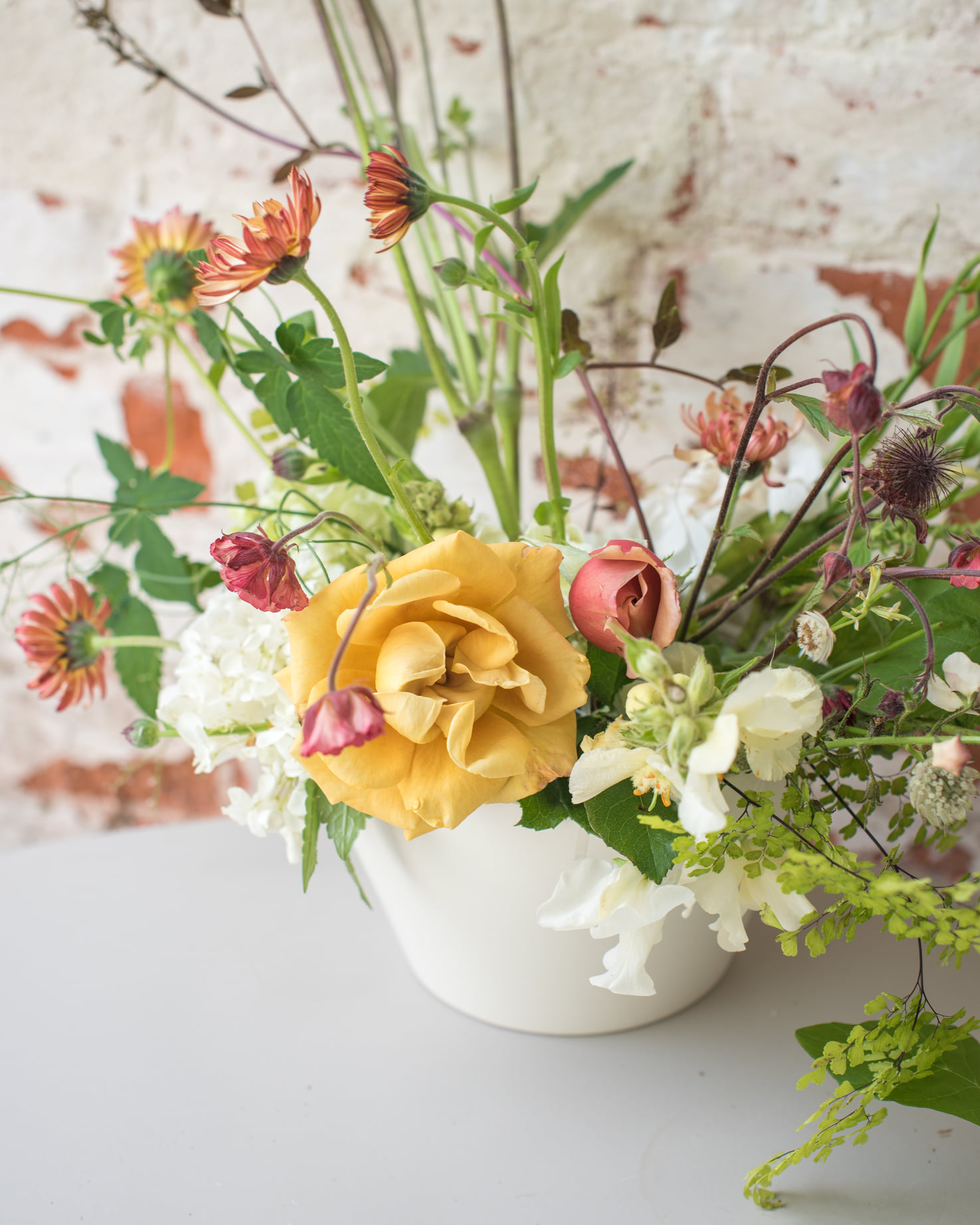 Close-up of an artisanal arrangement featuring a golden yellow rose, white hydrangea, and wild trailing stems in a ceramic vessel