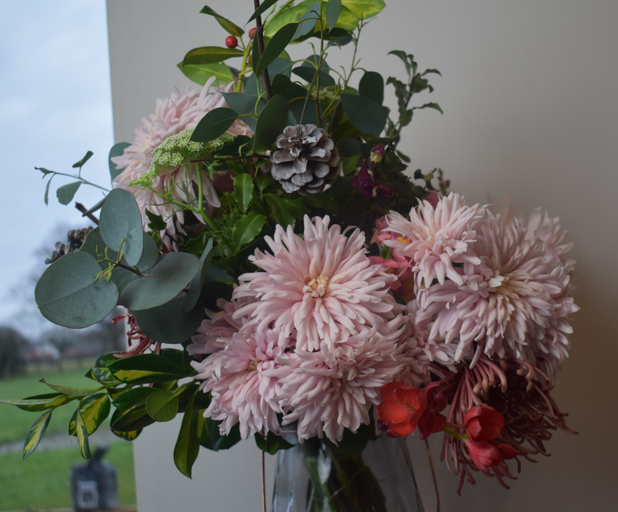 Lush arrangement of blush pink dahlias with eucalyptus, red accents, and a frosted pinecone