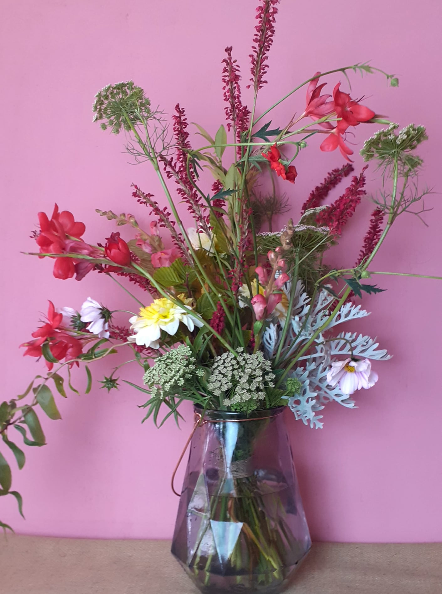 Vibrant mixed bouquet with red flowers, purple heather, and Queen Anne's lace in a glass vase against a pink wall