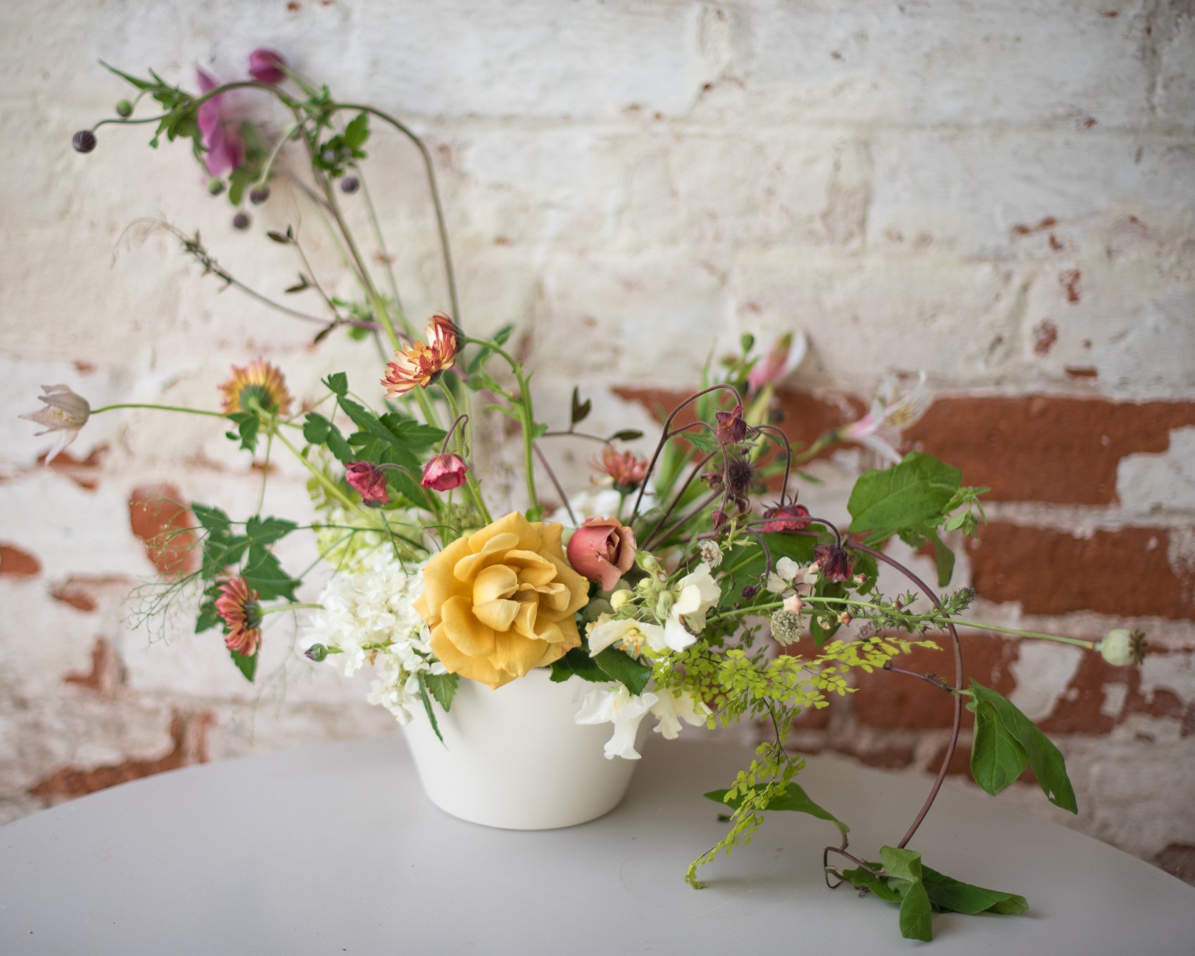 Artisanal flower arrangement with a yellow garden rose and wildflowers in a white ceramic vessel against a rustic brick wall